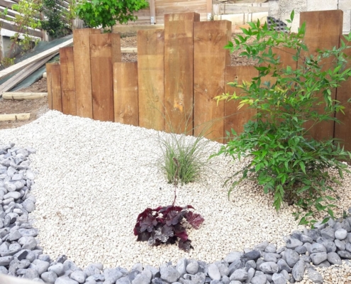 Photo représentant l'activité d'aménagement paysager que réalise OASURE. Elle est illustré par une création de jardin sec avec des graviers blancs au sol, un chemin en galets gris, des arbustes plantés sur les cotés et un contour avec des planches de bois de différentes tailles.