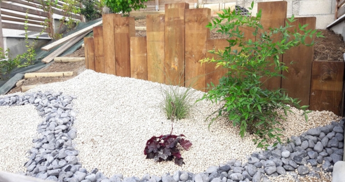 Photo représentant l'activité d'aménagement paysager que réalise OASURE. Elle est illustré par une création de jardin sec avec des graviers blancs au sol, un chemin en galets gris, des arbustes plantés sur les cotés et un contour avec des planches de bois de différentes tailles.