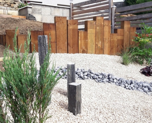 Photo représentant l'activité d'aménagement paysager que réalise OASURE. Elle est illustré par une création de jardin sec avec des graviers blancs au sol, un chemin en galets gris, des arbustes plantés sur les cotés et un contour avec des planches de bois de différentes tailles.