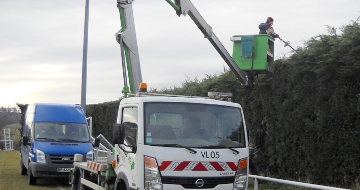 Photo représentant l'activité d'entretien des espaces verts d'OASURE. On observe un salarié suspendu sur une nacelle pour tailler le haut d'une grande haie.